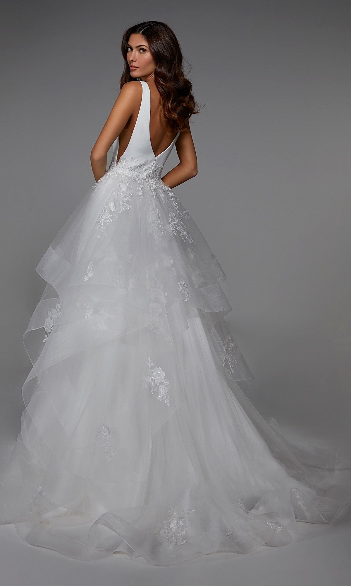 A woman stands against a plain gray background wearing the Alyce 7040, a long formal sleeveless and backless white tulle dress featuring layered tulle and floral lace details.